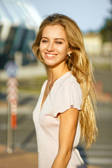 Positive blonde woman with long hair posing at the background of street in sun light