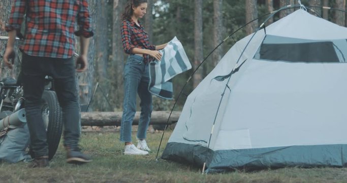 Hiker camping in forest. Handsome young man tourist puts hiking backpack in tent. Girl puts plaid on hiking mat. Countryside lifestyle scene. 4K video shooting by handheld gimbal