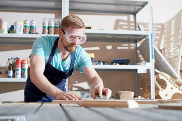 Young bearded carpenter wearing apron and glasses sanding wooden parts at the table