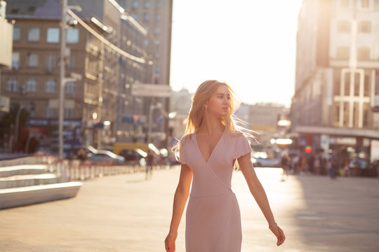 Amazing blonde woman in pink dress walking down the street in soft evening light. Space for text