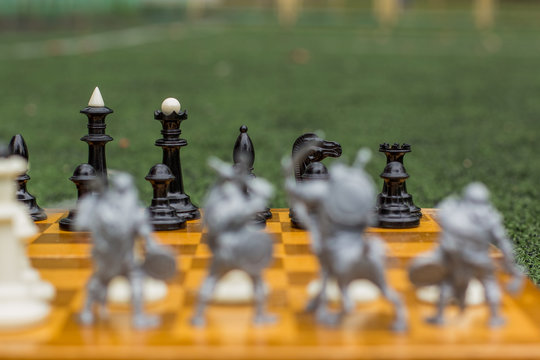 Soft Focus Classic Chess Game Concept On Wooden Yellow Desk Quadratic Marking And Unfocused Silhouettes Of Toy Soldiers Back To Camera On Green Grass Background