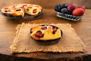 Piece of plum cake laid on a round plate, linen cloth napkin, ripe plums in the background, wooden background.