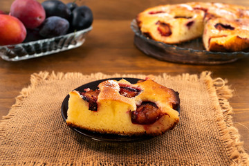 A cut piece of plum cake baked in the oven, ripe plums in a plate in the background, a linen cloth napkin, a wooden background.