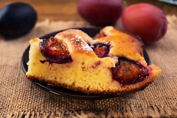 A portion of baked pie with plums on a plate, ripe plums blue and purple on a napkin of linen fabric.