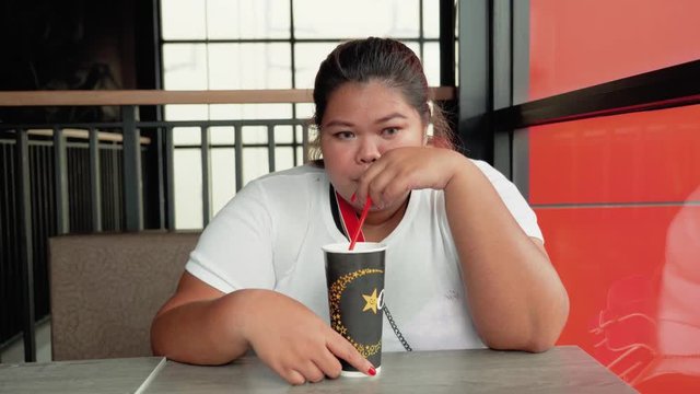 JAKARTA, Indonesia - September 04, 2018: Overweight young woman drinks a cup of soft drink in the cafe. Shot in 4k resolution