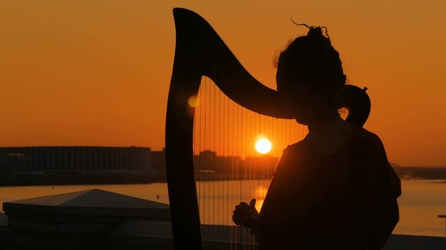 Portrait of unrecognizable woman silhouette playing harp on city embankment at sunset. Music, leisure and culture concept