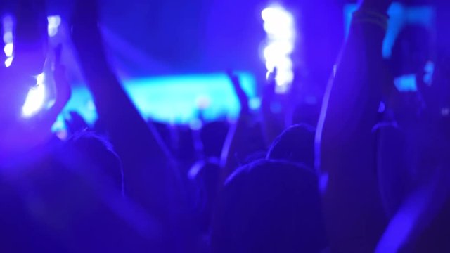 A slow motion of a clapping crowd on an open air concert show. It is a dark evening outside and everything is filled with blue illumination from the bright stage
