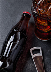 Bottle of cola soda drink and glass with ice cubes next to opener on black stone background.