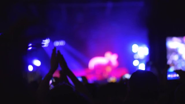 A slow motion of a cheering crowd on an open air concert show. It is evening outside and the blurred stage is bright and colorful, illuminated with lots of lights. Clapping hands are everywhere and