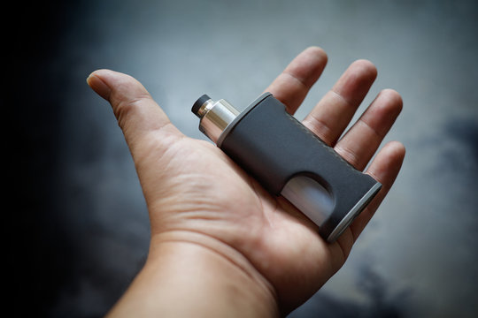 high end unregulated bottom feed squonk box mods with rebuildable dripping atomizer in hand on dark grey texture background, vaping device, vape gear, vaporizer equipment, selective focus