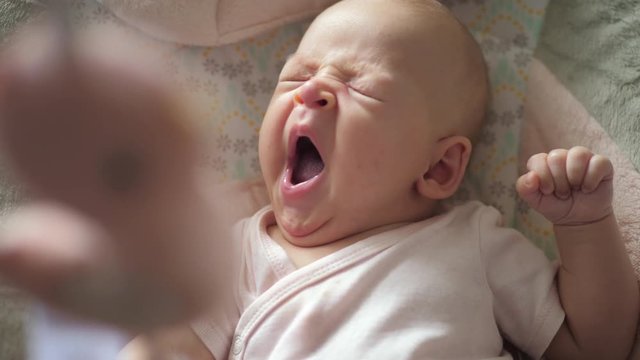 A two months old baby girl is lying on a back on patterned bed linen. She is yawning so hard that she is squinting and that is looking funny and adorable