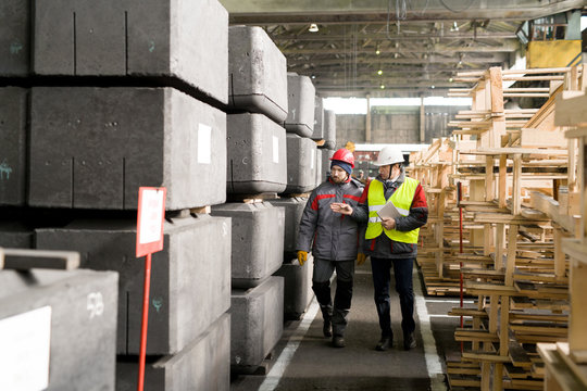 Full length portrait of mature foreman wearing hardhat  discussing manufacturing with worker while walking by concrete blocks in workshop
