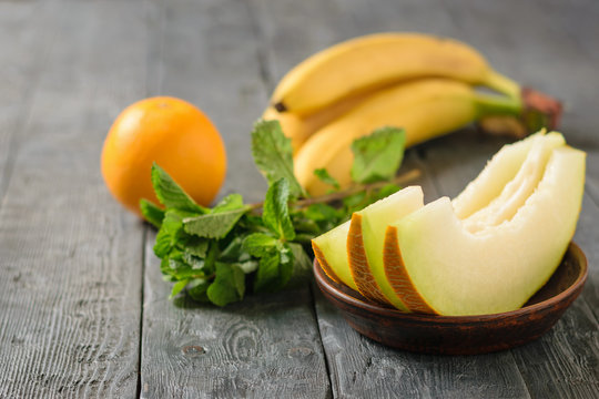 Ripe melon pieces on a clay bowl, banana, mint and orange on a wooden table.