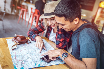 Young casual woman and man reading paper map and planning route while sitting at table in cafe