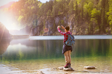 Back view of young man takes pictures by cellphone with a beautiful landscape and sun flare as lens...