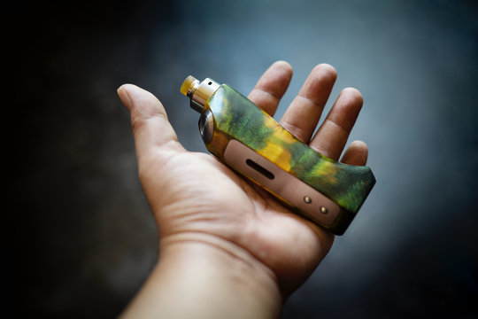 high end yellow green stabilized wood box mods with rebuildable dripping atomizer in hand on dark grey texture background, vaping device, vape gear, vaporizer equipment, selective focus