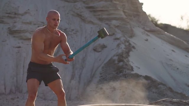 Bald man strongman hits a hammer on a huge wheel in the sandy mountains in slow motion. strength and endurance training for wrestlers.