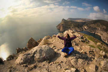 Beautiful young happy carefree woman traveler sits on top of a hill with a stunning amazing sea view, with arms outstretched