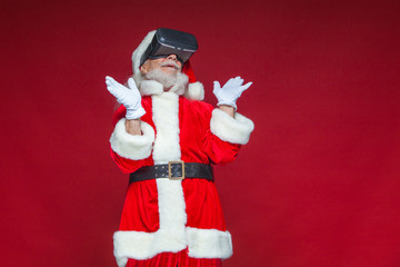 Fototapeta na wymiar Christmas. Santa Claus in black virtual reality glasses makes gestures with his hands. Surprise, emotion. New technology. Isolated on red background.