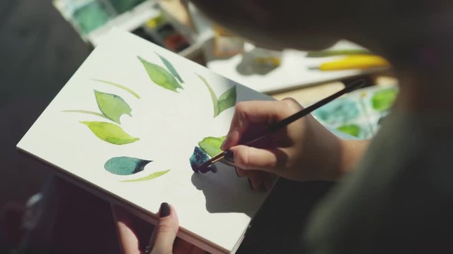 Artist made a sketch in pencil on a sheet of album and paints, painting leaves of plants. Create a floral print, start drawing. A female hand is holding a brush and doing smears. Filling with color.