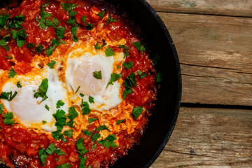 Homemade breakfast shakshuka - fried eggs, onion, bell pepper, tomatoes and parsley in cast-iron frying pan on rustic wooden table. Jewish cuisine. Top view
