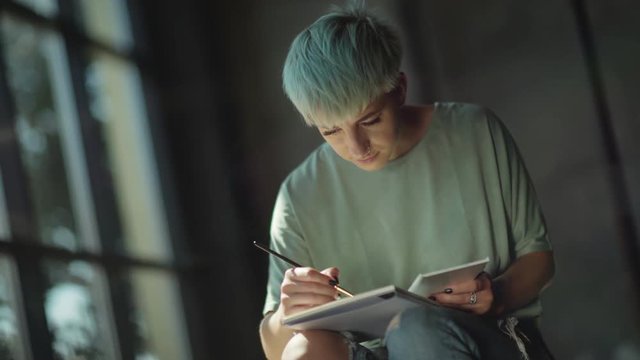 Young woman with a short haircut, colored hair, piercer holds on her knees an album for drawing. Artist paints with paintbrush, smiling in the process. Inspiration. An artist with a bright appearance.