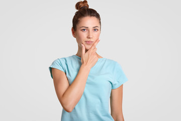 Thoughtful puzzled woman with hesitant expression, holds chin, dressed in casual clothes, ponders...