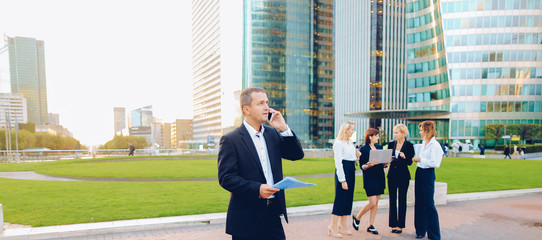 Businessman speaking by smartphone outside, keeping blue document case with employees in...
