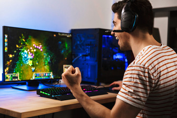 Portrait of delighted young guy playing video games on computer, wearing headphones and using backlit colorful keyboard