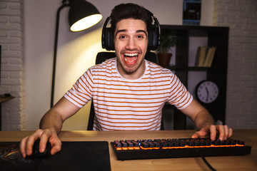 Portrait of caucasian guy wearing headphones smiling while looking on computer, and using backlit colorful keyboard