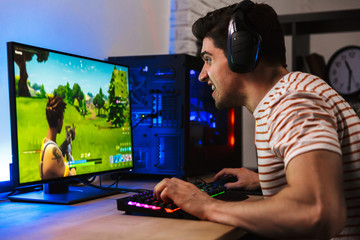 Portrait of caucasian gamer guy looking at screen while playing video games on computer, wearing headphones and using backlit colorful keyboard - obrazy, fototapety, plakaty