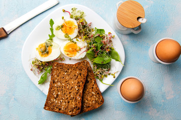 Zdrowe śniadanie - Jajka gotowane na twardo, świeże kiełki rzodkiewki, rukola z sosem ziołowym i kromki pełnoziarnistego ciemnego chleba na błękitnym tle. 