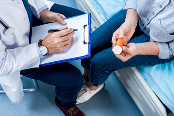 Obraz premium cropped image of mature male doctor writing in clipboard near female patient with pills in hospital room