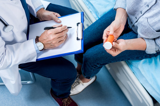 cropped image of mature male doctor writing in clipboard near female patient  with pills in hospital room