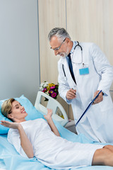 smiling male doctor with stethoscope over neck holding clipboard and talking to female patient in hospital room