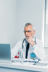 serious middle aged male doctor using laptop at table in office