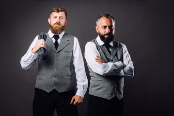 Two bearded men in strict suits standing one near one and looking on camera on black