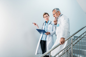 adult female doctor showing clipboard to thoughtful male colleague on staircase in hospital