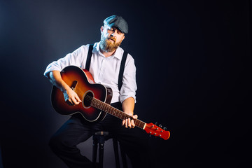 Fototapeta premium A bearded man in suit and hat sitting and playing on the acoustic guitar. Studio portrait