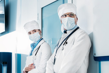 male and female doctors in medical masks standing and looking away in hospital corridor