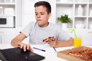 Young teenager boy working on a project having a bite of pizza at the kitchen desk
