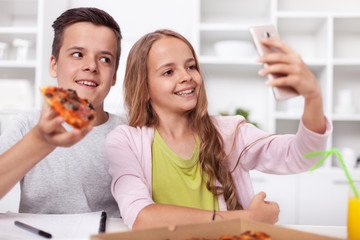 Young teenager boy and girl taking a selfie - eating pizza in the kitchen