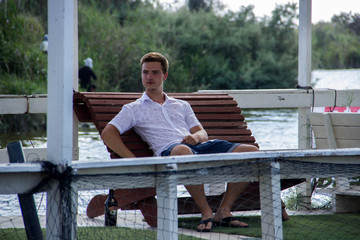 A young attractive man in a white shirt sits on a floating restaurant in the background of a pond