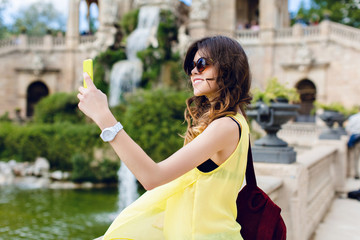 Brunette girl is making selfie-portrait on sightseeing background. She wears yellow T-shirt,...