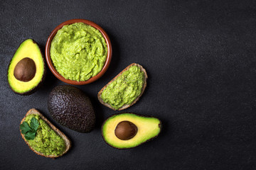 Traditional Mexican Dip Sauce Guacamole in a bowl with bread toasts,  whole and cut half avocado  on dark background. Top view. Copy space. - obrazy, fototapety, plakaty