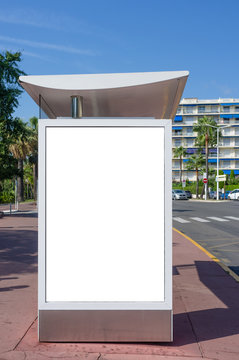 Vertical blank white billboard at bus stop on city street. In the background buildings and road. Mock up. Poster on street next to roadway. Sunny summer day. Cannes, France