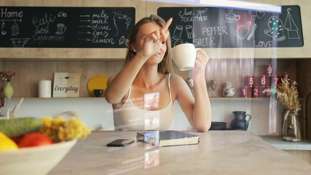 Woman has morning coffee while browsing internet social network via futuristic hologram interface panel sitting on the kitchen table, future transparent visual touchscreen in use