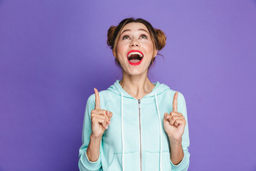 Portrait of stylish beautiful girl with two buns pointing fingers upward at copyspace with open mouth, isolated over violet background in studio