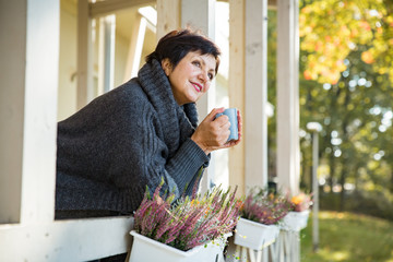 Mature attractive woman standing on Cozy wooden terrace with cup of hot coffee wrapped up in knitted warm sweater, happy smile. Lake house in autumn, yellow and red leaves on trees. Sunny day