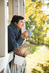 Mature attractive woman standing on Cozy wooden terrace with cup of hot coffee wrapped up in knitted warm sweater, happy smile. Lake house in autumn, yellow and red leaves on trees. Sunny day